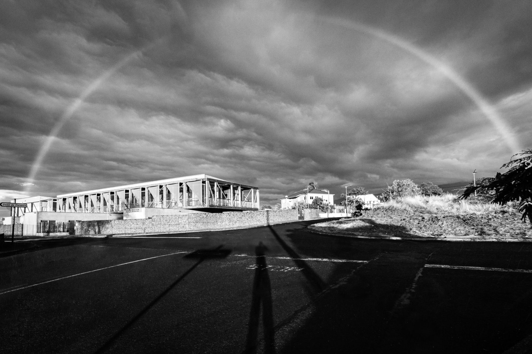 Photographie noir et blanc de la Direction de la Mer du Sud Océan Indien dont l' architecte se nomme Olivier Brabant. C'est une photographie prise au grand angle montrant l'étifice sous une couche de nuages et un arc en ciel.